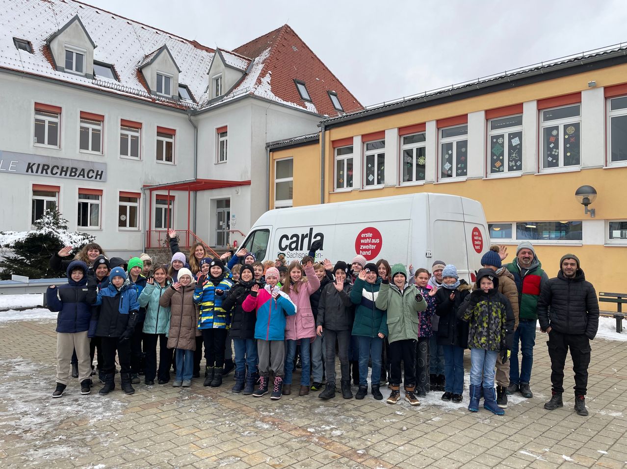 Eine Gruppe von Kindern und Erwachsenen in Winterkleidung posiert für ein Foto vor einem Gebäude mit einem weißen Lieferwagen mit dem Wort Carla.