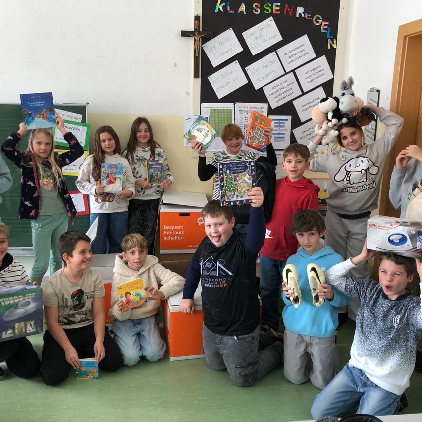 Mehrere Kinder in einem Klassenzimmer halten Bücher und Spielzeug und posieren für ein Foto, einige halten Kuscheltiere und Kisten. Ein Bulletin Board im Hintergrund mit Papieren und einem Kreuz.