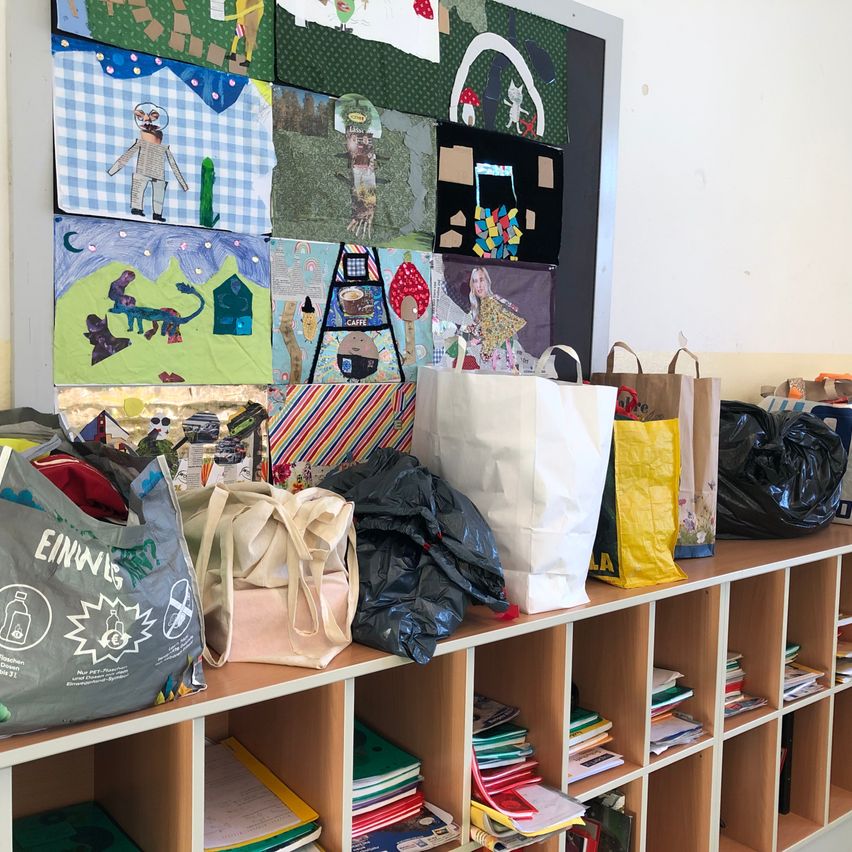 Ein Klassenzimmer zeigt verschiedene Kunstwerke an der Wand. Taschen, Bücher und andere Gegenstände sind unter den Regalen aufbewahrt.
