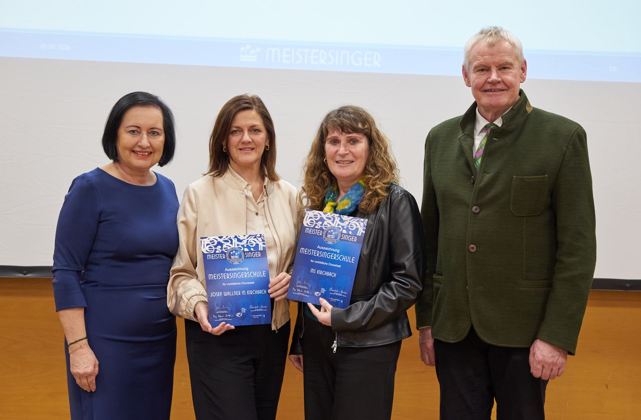 Vier Menschen stehen zusammen, halten Auszeichnungen in der Hand, lächeln und stehen vor einem Bildschirm mit dem Text Meistersinger. Sie scheinen eine Anerkennung zu erhalten.