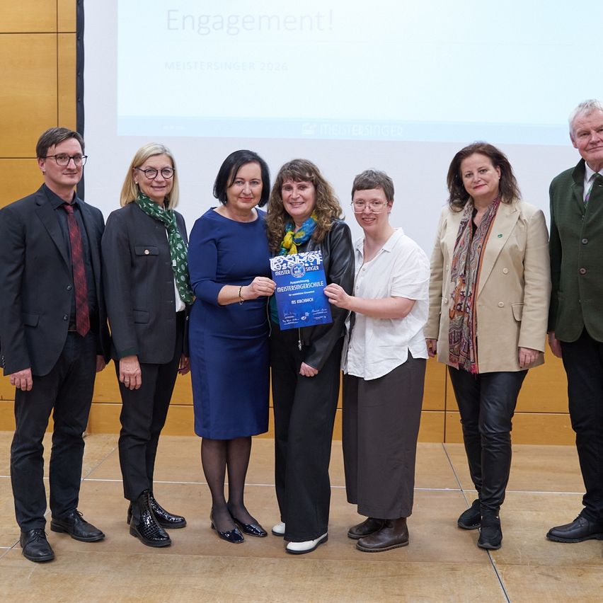 Eine Gruppe von Menschen steht vor einer Projektionsleinwand mit dem Text Engagement! Meistersingers 2020. Sie posieren für ein Foto mit einem blauen Zertifikat.