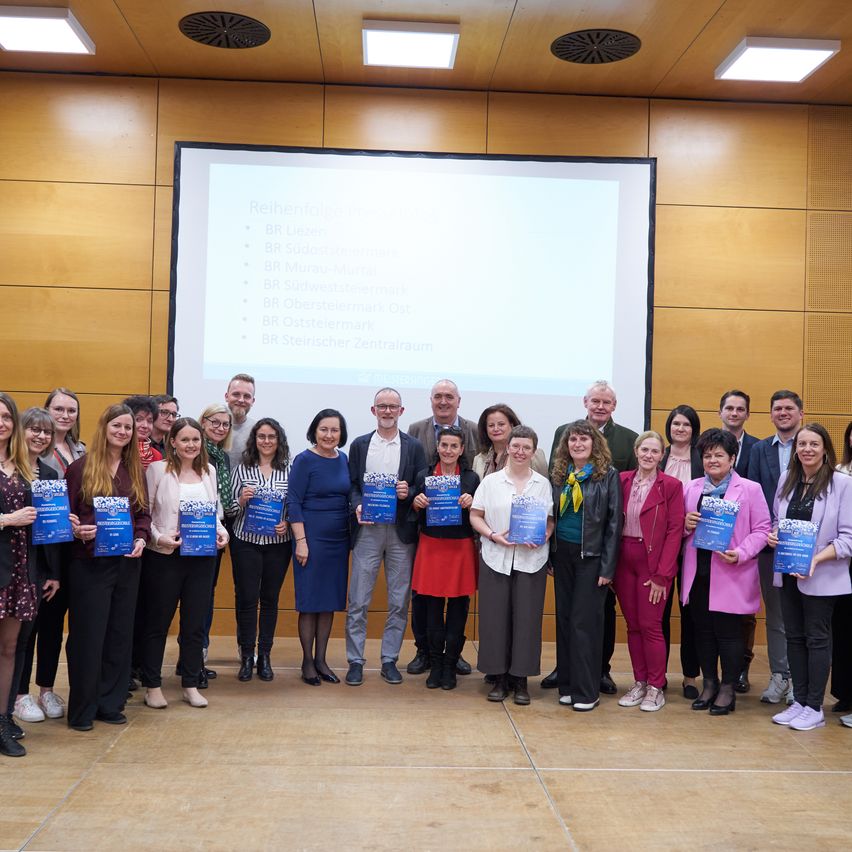 Eine Gruppe von Menschen steht vor einer Projektionsleinwand, hält Urkunden in der Hand und lächelt für ein Foto.