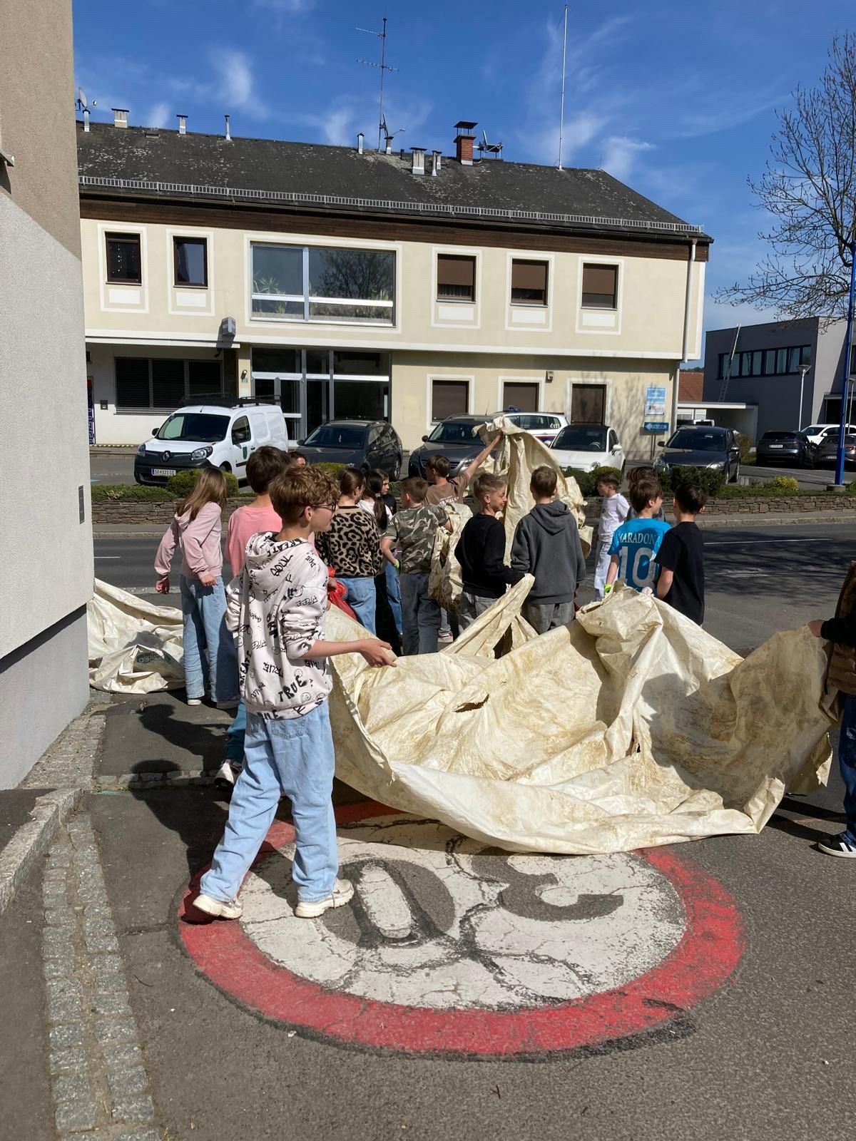 A group of children, some in hoodies and sneakers, are gathered around a large white cloth on the road. They are outside a building with glass windows. Cars are parked in front of the building.