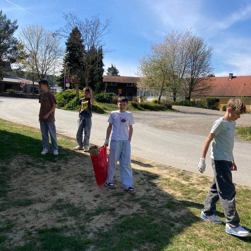 Four boys stand in a grassy area, one holding a red bag, while another gestures towards the road. The weather is sunny.
