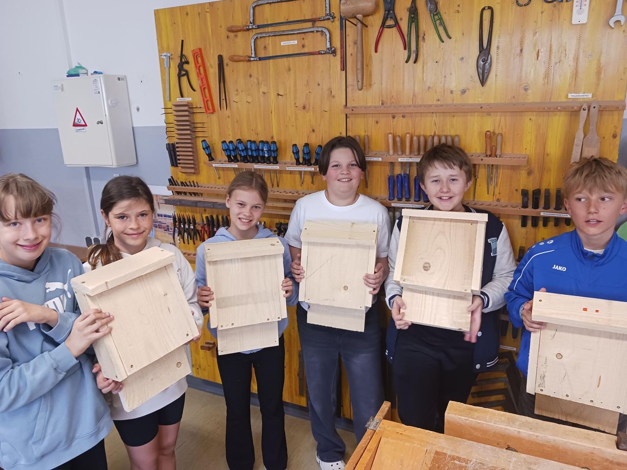 Fünf Kinder halten Holzvogelhäuschen vor einer mit Werkzeug gefüllten Wand hoch. Sie lächeln für die Kamera.