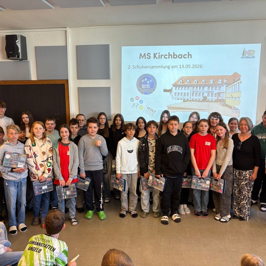 Schüler und Lehrer der MS Kirchbach Schule versammeln sich für eine Versammlung, stehen in Reihen mit Zertifikaten und einer großen Leinwand, die Datum und Logo der Veranstaltung anzeigt.