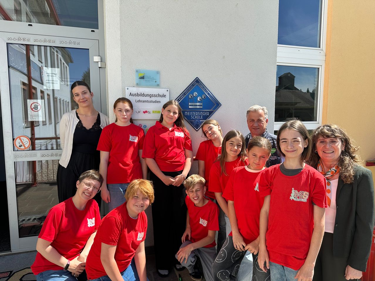 Eine Gruppe von Schülern in roten Shirts steht vor einem Gebäude mit einem Schild, das 'Ausbildungsschule Lehramtsstudien' besagt. Sie lächeln und posieren für ein Foto.