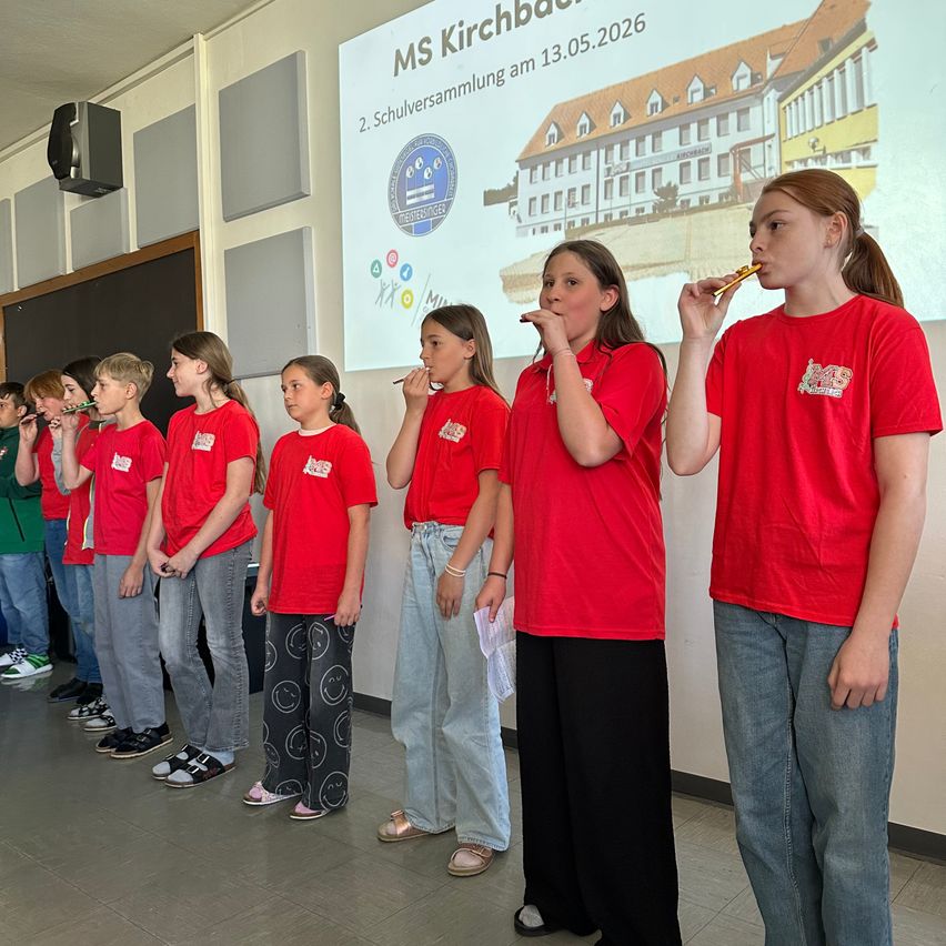 Schüler in roten Shirts stehen in einer Reihe und spielen Blockflöten in einem Klassenzimmer. Eine Projektionsleinwand zeigt ein Datum und ein Schullogo.