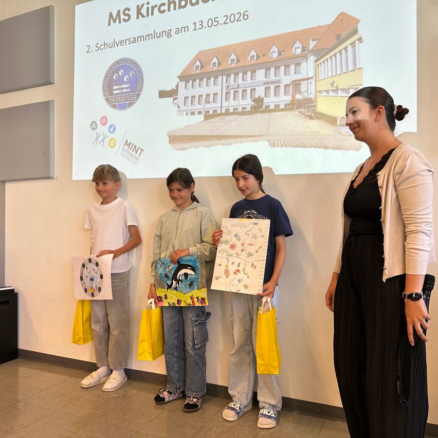 Drei Kinder stehen vor einer Leinwand mit einem Gebäudeabbild. Sie halten jeweils einen Papierbeutel und eine Zeichnung. Eine Frau in formeller Kleidung steht neben ihnen.