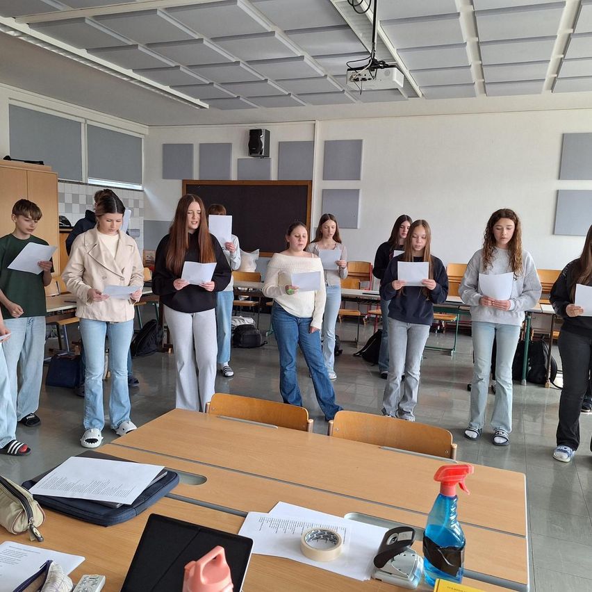 Eine Gruppe von Schülern in einem Klassenzimmer hält Papiere in der Hand und steht in einer Reihe, wahrscheinlich im Proben für eine Aufführung. Der Raum ist mit Tischen, Stühlen und einer Tafel ausgestattet.