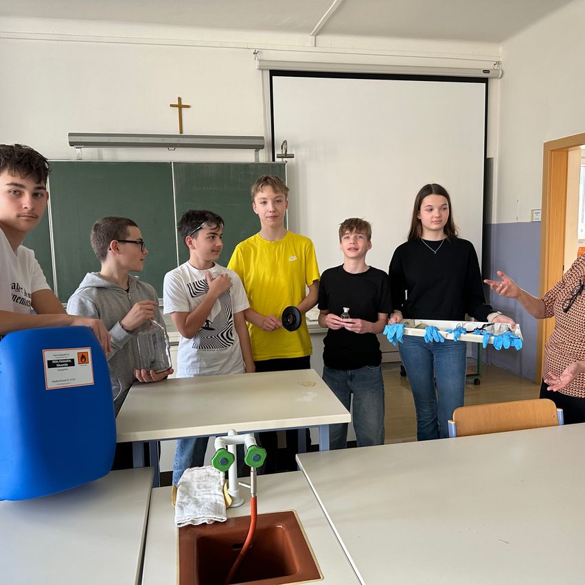 Eine Gruppe von Schülern in einem Labor, eine Frau unterrichtet sie vor einem Waschbecken. Hinter ihnen ist ein Klassenzimmer mit einer Tafel und einem Kreuz an der Wand.