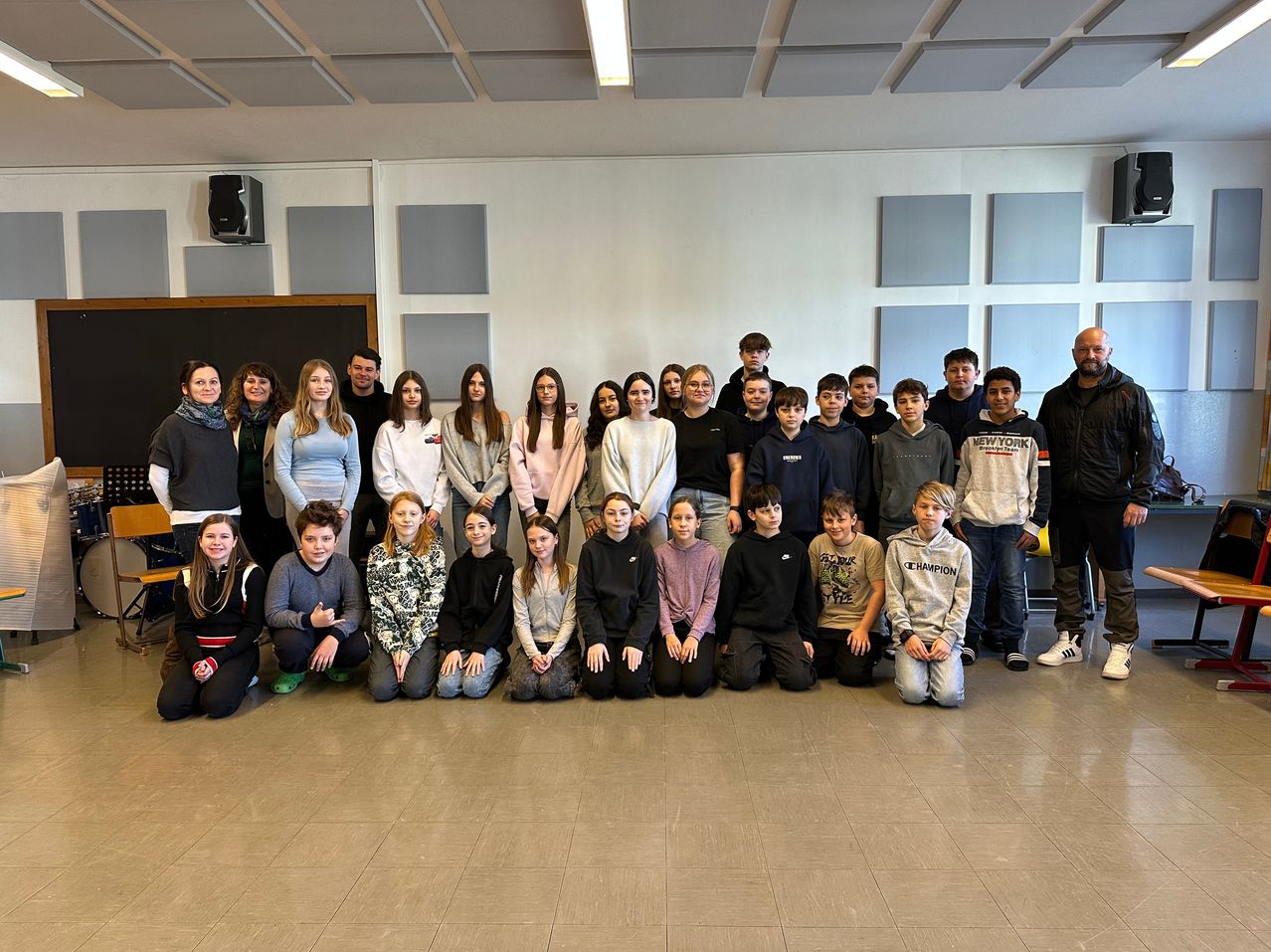 Eine Gruppe von Schülern und Lehrern posiert für ein Foto im Klassenzimmer. Die Schüler sitzen und stehen vor einer Wand mit einer Tafel.