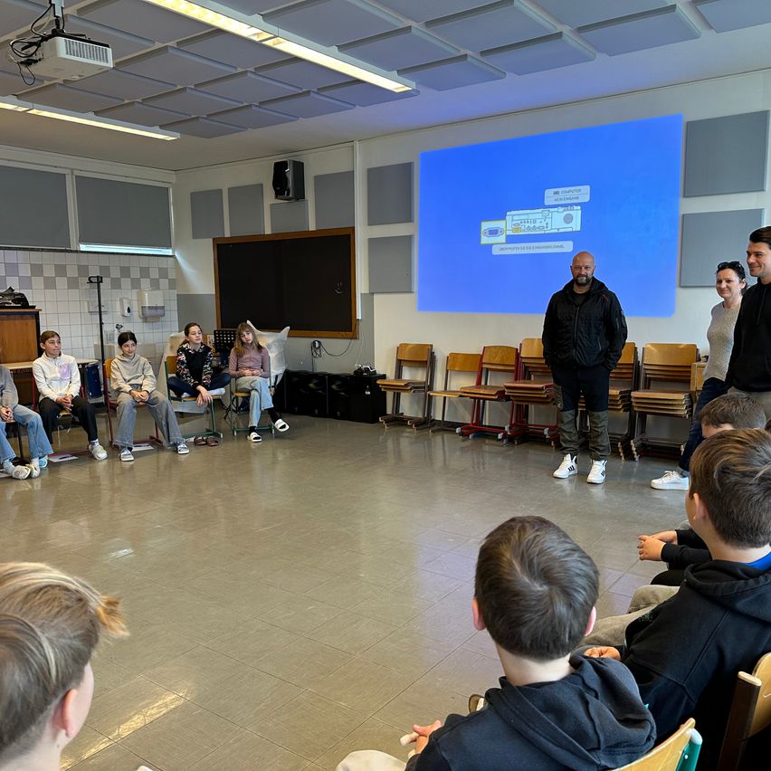 Eine Gruppe von Menschen, einschließlich eines Mannes, der vor einem Projektor steht, versammelt sich in einem Raum. Sie scheinen einer Präsentation zuzuhören.