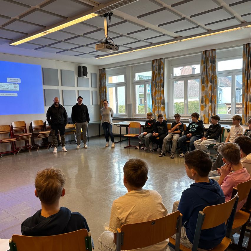 Eine Gruppe von Menschen in einem Klassenzimmer. Zwei Erwachsene stehen vor einer Leinwand, während mehrere Schüler auf Stühlen um sie herum sitzen.