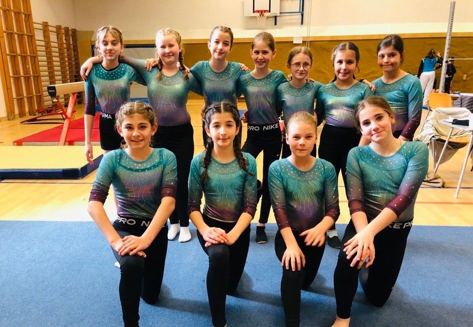 Eine Gruppe junger Mädchen in einheitlichen Outfits posiert für ein Foto in einer Turnhalle, mit einem Basketballkorb im Hintergrund.