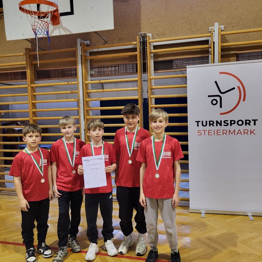 Fünf Jungen in roten Trikots posieren mit Medaillen und einem Zertifikat in einer Turnhalle. Ein Basketballkorb ist hinter ihnen sichtbar.