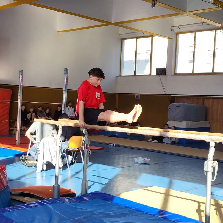 Ein junger Junge in einem roten T-Shirt führt eine Turnübung an der horizontalen Stange in einer Turnhalle aus. Zuschauer sitzen auf dem Boden, und Matten sind im Bereich platziert.