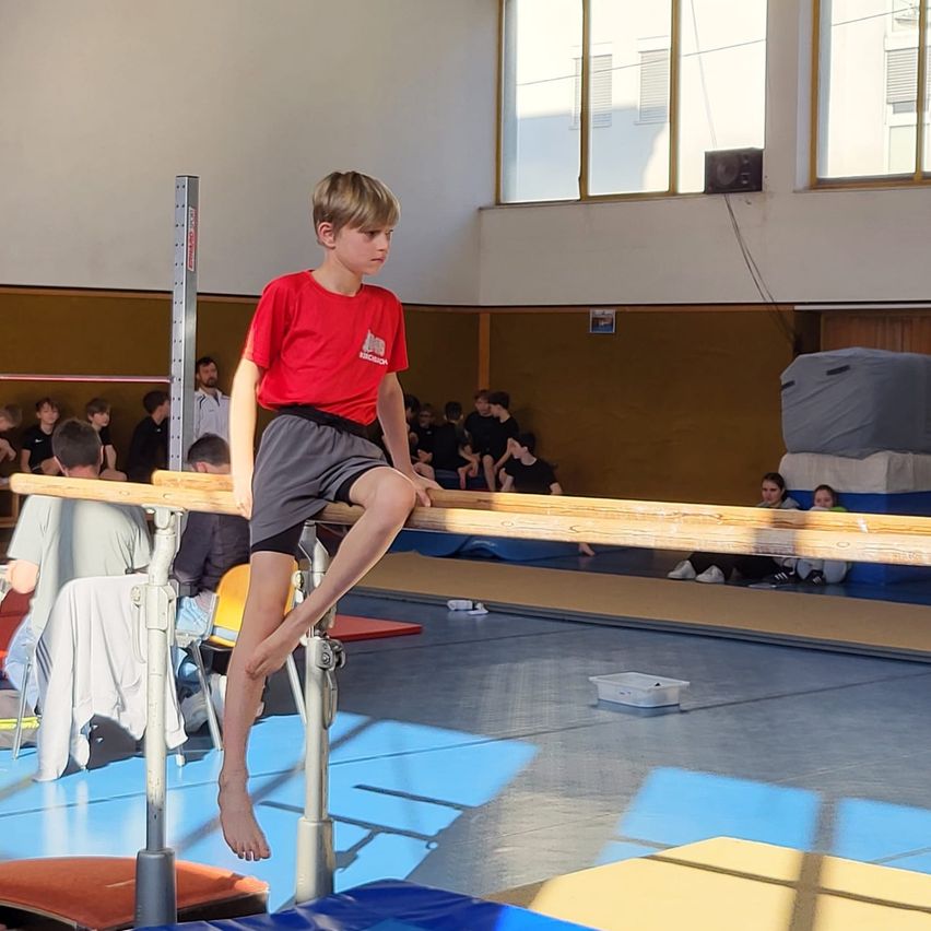 Ein junger Junge balanciert auf einer Stange in einem Fitnessstudio, trägt ein rotes T-Shirt und Shorts. Zuschauer sitzen an den Seiten, und Fitnessgeräte sind im Hintergrund sichtbar.