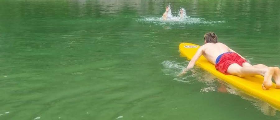 Ein Junge liegt auf einem gelben Surfbrett im grünen Wasser. Im Hintergrund springen zwei Personen von einem Felsen ins Wasser.