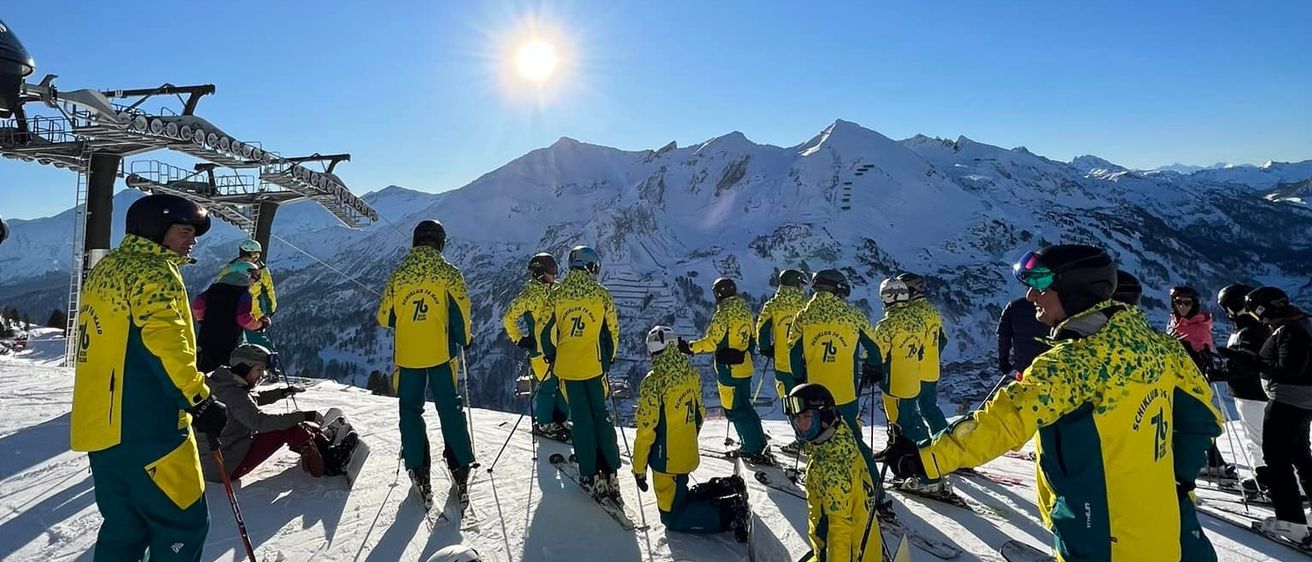 Eine Gruppe von Skifahrern in gelben und grünen Uniformen steht auf einem verschneiten Berg, eine Person kniet und andere stehen mit Skistöcken. Die Sonne scheint hell am Himmel.