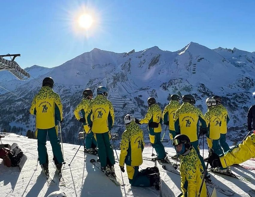 Eine Gruppe von Skifahrern in gelben und grünen Uniformen steht auf einem verschneiten Berg, eine Person kniet und andere stehen mit Skistöcken. Die Sonne scheint hell am Himmel.
