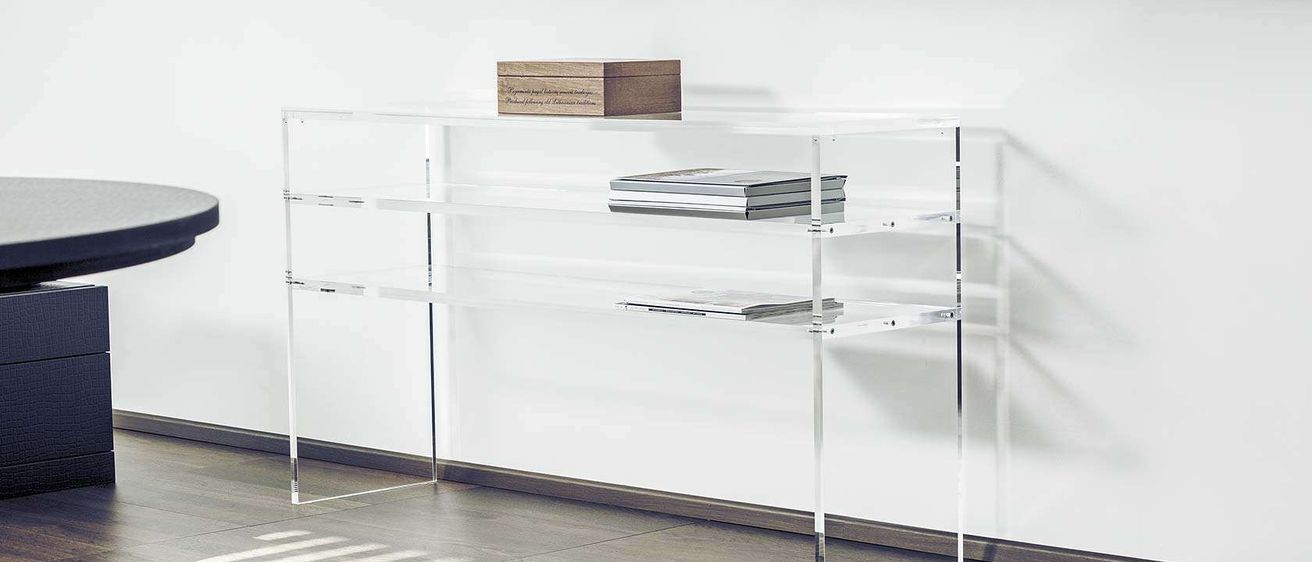 Ein klarer Acryl-Konsolentisch mit drei Regalen steht an einer weißen Wand. Auf dem obersten Regal befindet sich eine Holzkiste und auf den beiden unteren Regalen mehrere Bücher.