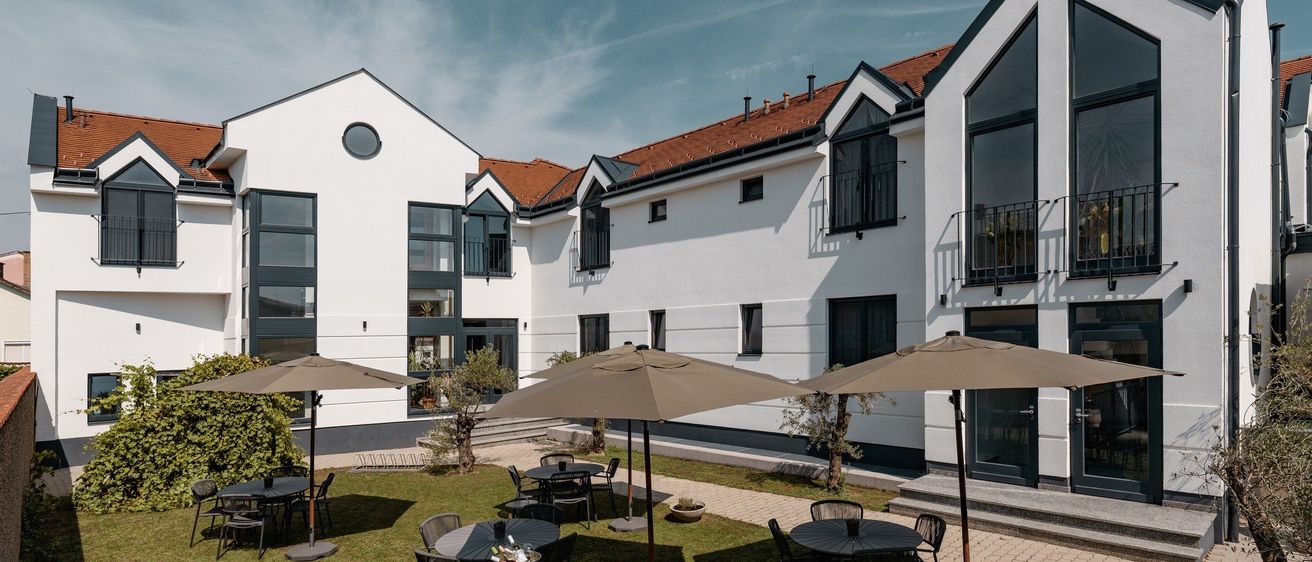 Ein modernes weißes Gebäude mit einem braunen Dach, mehreren Fenstern und Balkonen steht in einem grasigen Innenhof. Tische und Stühle sind unter Schirmen angeordnet, und ein Fass steht in der Mitte.