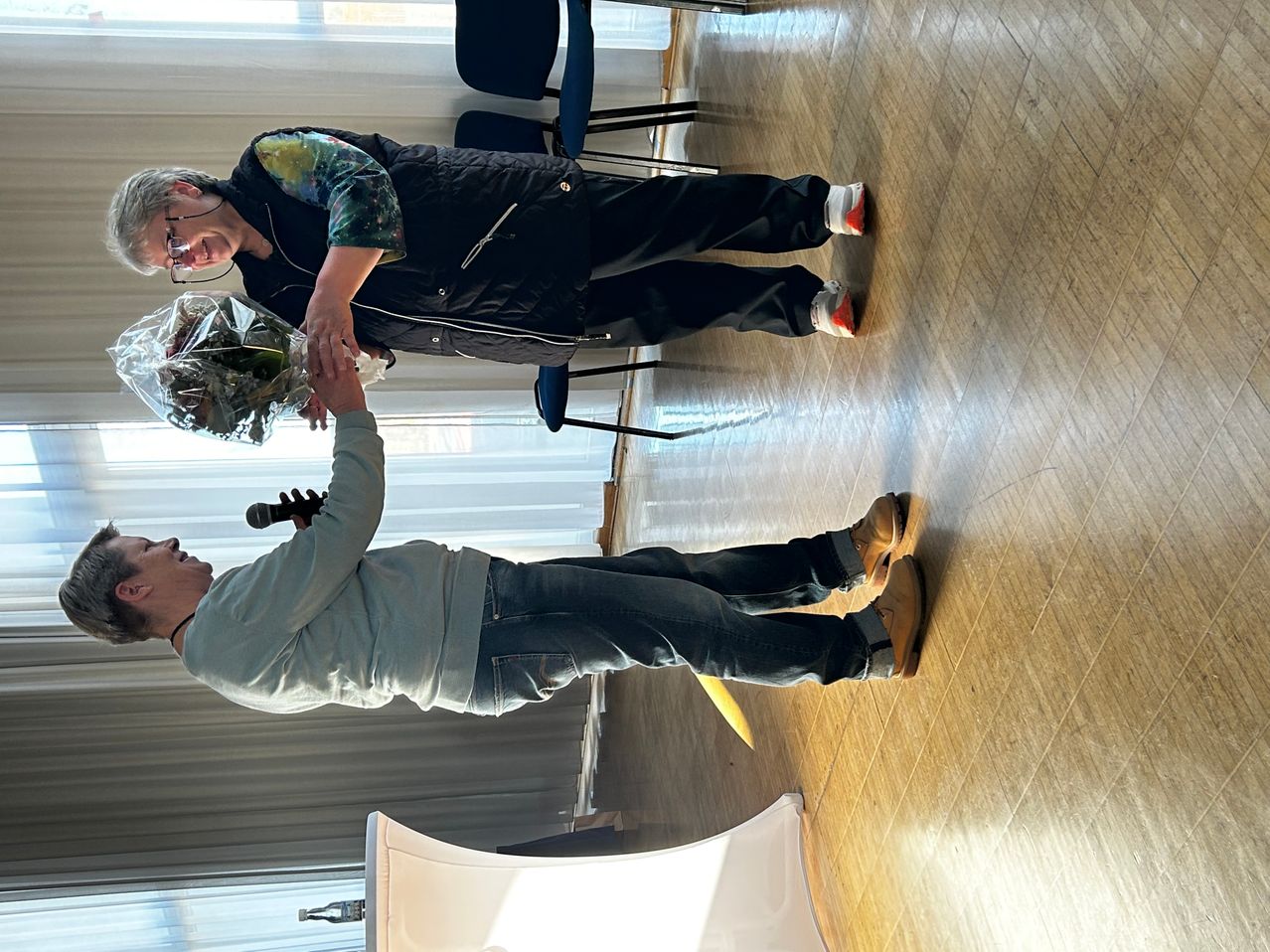 Ein Mann in einem grauen Pullover und Jeans verbeugt sich vor einer Frau, die einen Blumenstrauß hält. Die Frau trägt eine schwarze Jacke und Hose. Dahinter sind zwei blaue Stühle gestapelt. Der Boden ist aus Holz.