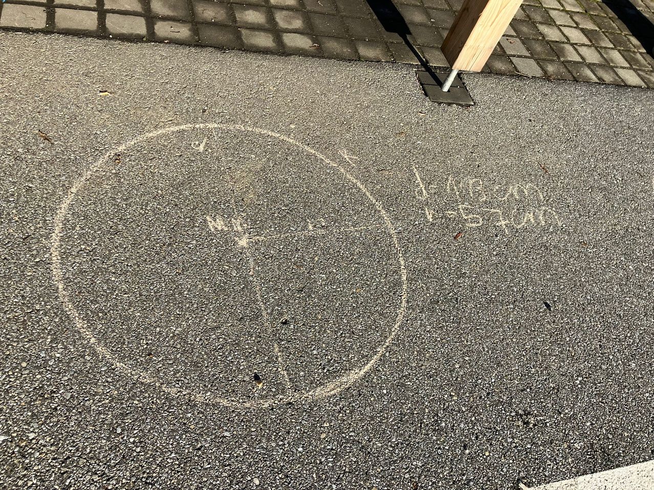 Ein Beton-Gehweg mit einer kreisförmigen Kreidezeichnung darauf, umgeben von einem Ziegelstein-Pflaster. Die Kreidezeichnung enthält die Worte 'HALLO' und 'VISION'. Ein Holzpfosten befindet sich in der Nähe.