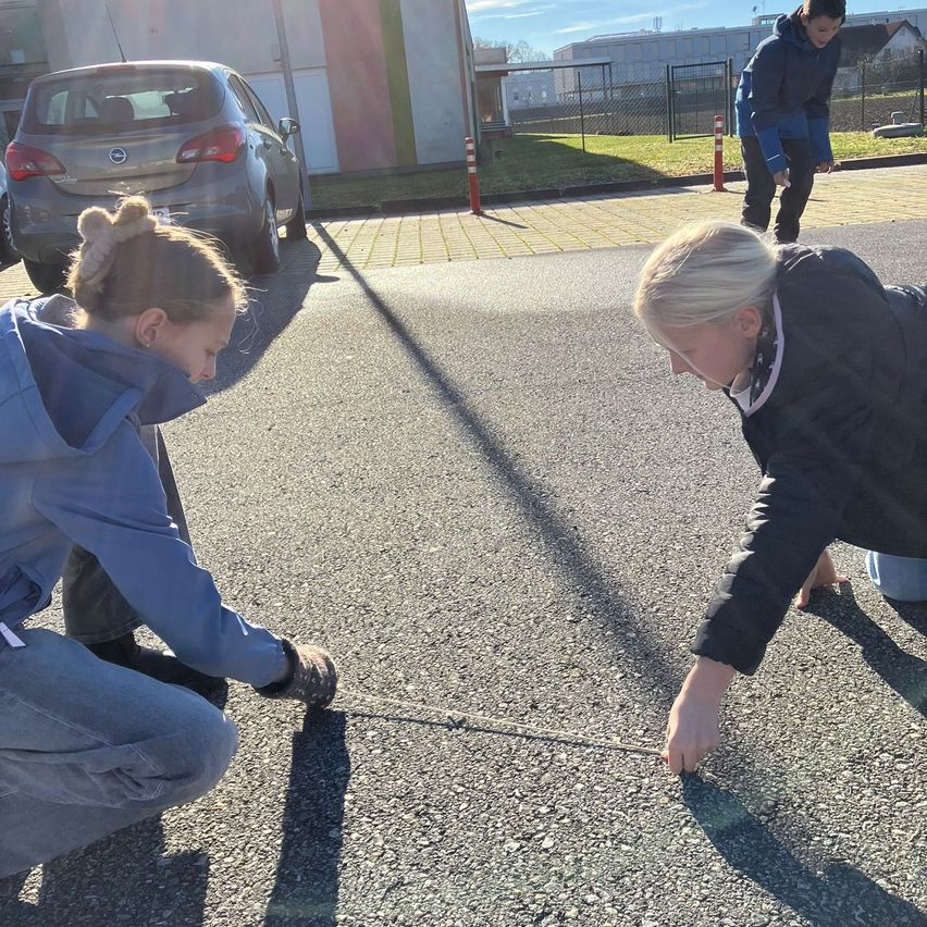 Zwei Frauen messen eine Entfernung auf einer Straße, eine kniet und die andere beugt sich vor. Ein Auto ist in der Nähe geparkt, und eine Person steht in der Ferne.