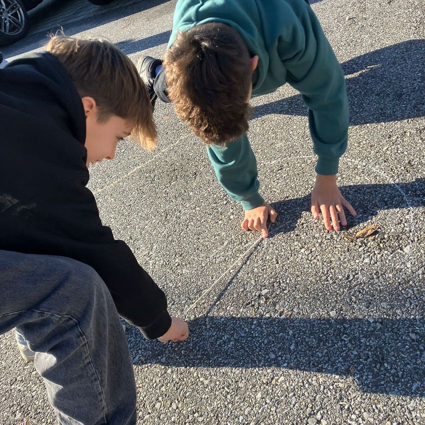 Zwei Jungen auf einer Straße, einer in Grün und einer in Schwarz, untersuchen etwas am Boden. Der Junge in Grün beugt sich nach unten, seine Hände sind auf dem Boden.