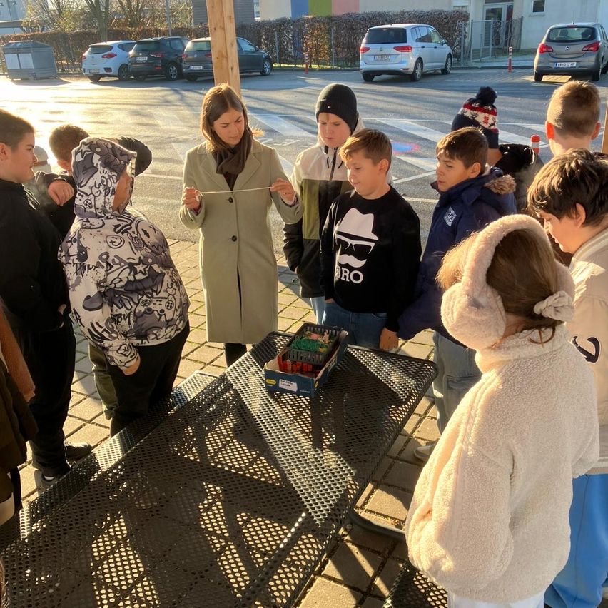 Eine Gruppe von Kindern und einer erwachsenen Frau versammelt sich um einen Tisch im Freien, auf dem eine Box platziert ist. Die erwachsene Frau hält eine Schnur und erklärt möglicherweise etwas den Kindern.