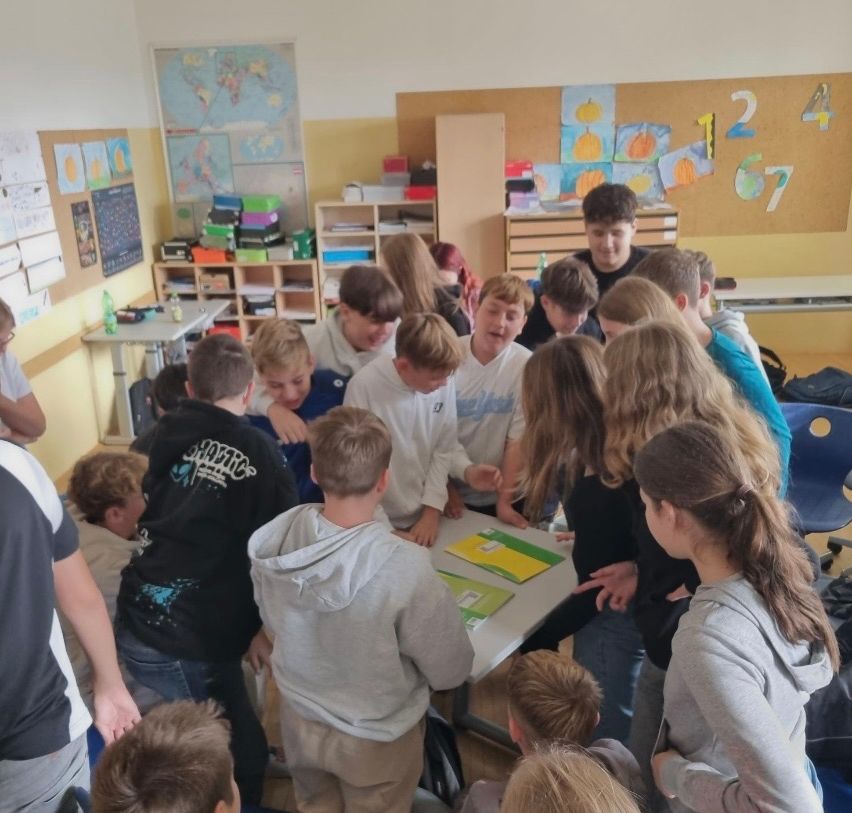 Eine Gruppe von Kindern und Erwachsenen versammelt sich um einen Tisch in einem Klassenzimmer. Sie schauen sich Papiere an und diskutieren etwas. An der Wand hängt eine Karte.