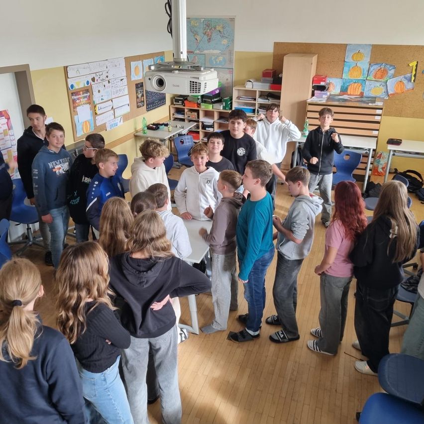 Ein Klassenzimmer mit Schülern, einige stehen, andere sitzen, mit einem Projektor an der Deckenmontage und verschiedenen Dekorationen an den Wänden.