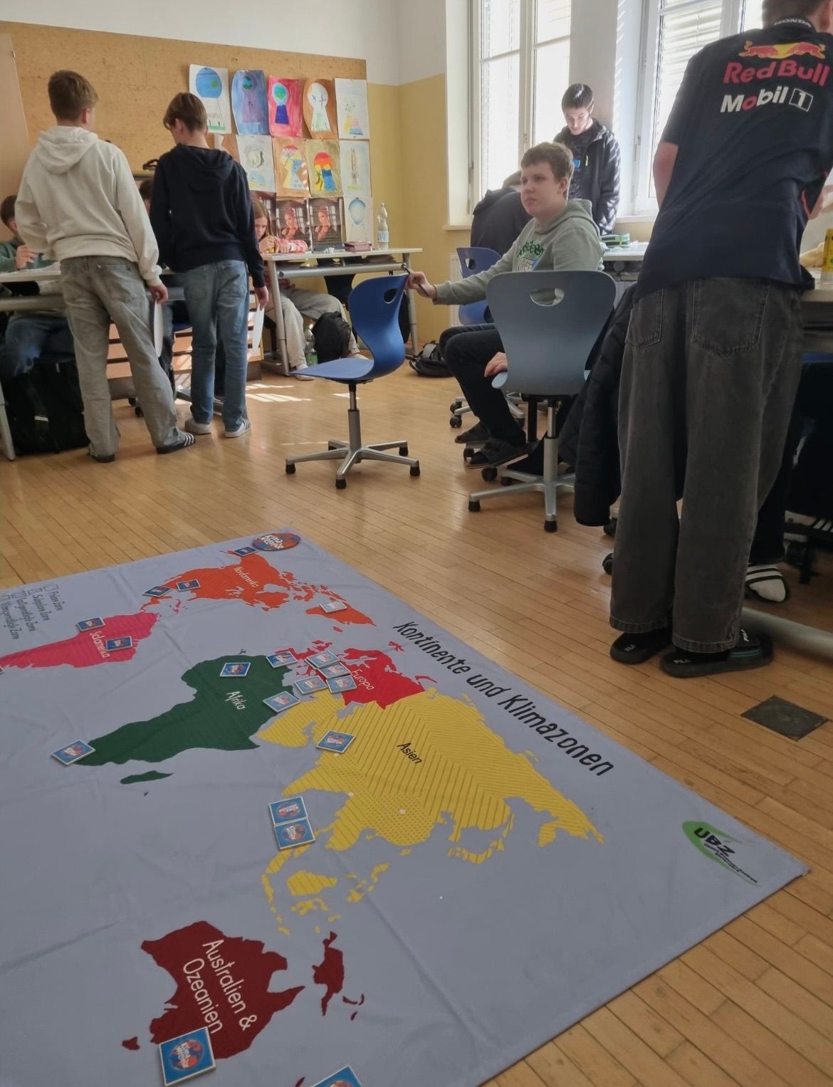 Schüler stehen und sitzen um eine große Weltkarte herum. Sie spielen ein Spiel, das die Kontinente und Klimazonen betrifft.