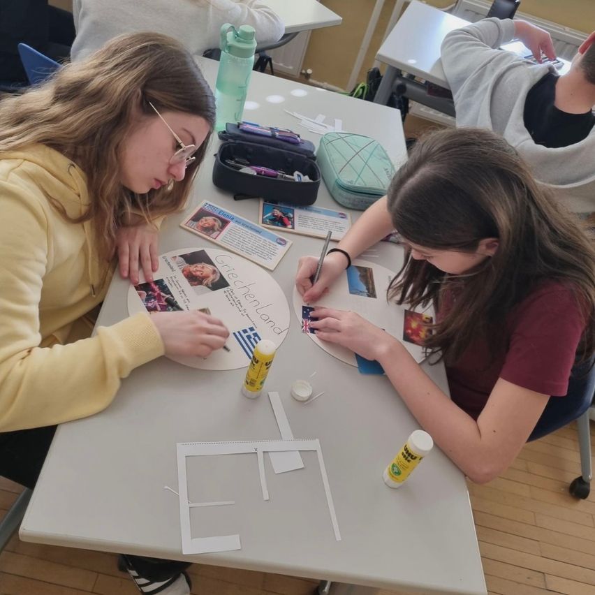 Zwei Schülerinnen sitzen an einem Tisch und arbeiten an einem Projekt. Sie haben Materialien wie Kleber, Papier und Flaggen. Eine schreibt auf ein Papier, das Griechenland sagt.