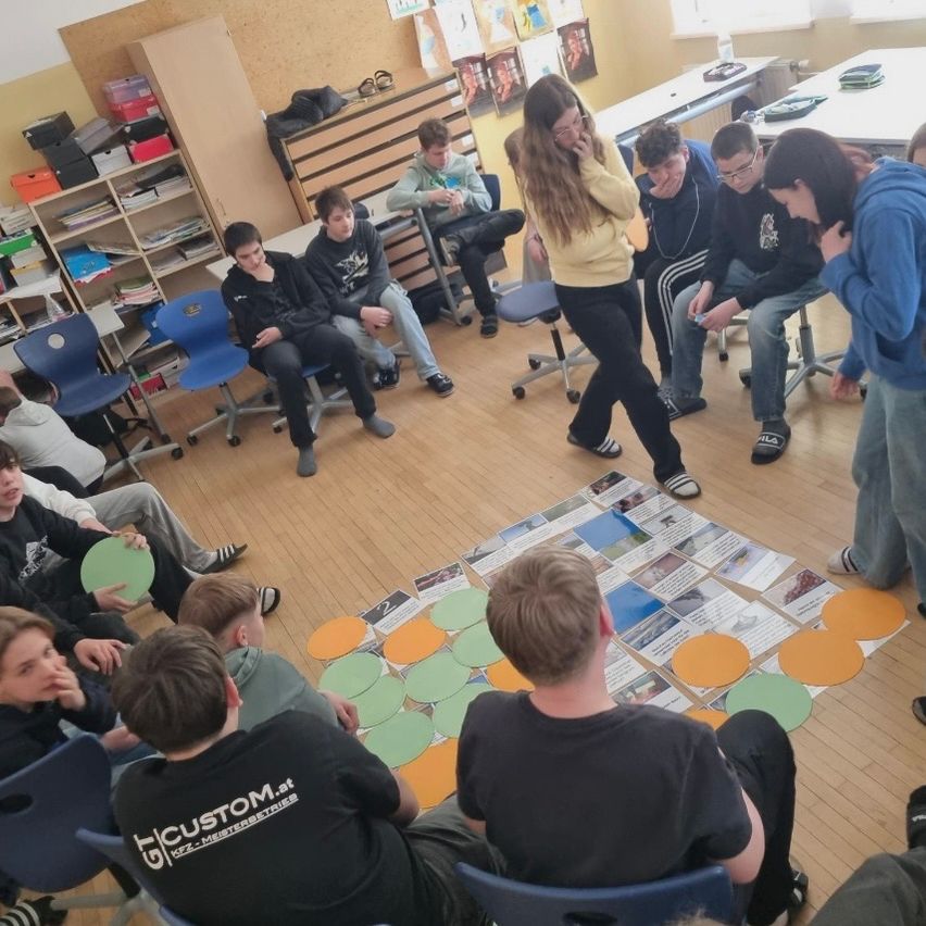 Ein Klassenzimmer mit einer Gruppe von Schülern, die an einer Gruppenaktivität teilnehmen. Eine Frau steht in der Mitte mit einem Brett. Die Schüler sitzen im Kreis auf dem Boden.