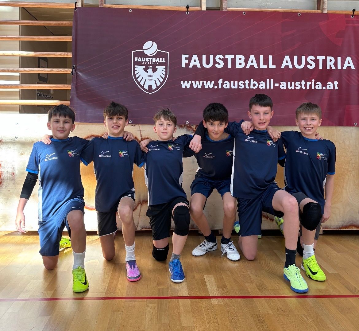 Sechs junge Jungen posieren vor einem roten Banner mit dem Text 'Faustball Austria' und einer Website. Sie tragen blaue Trikots mit weißen Streifen.