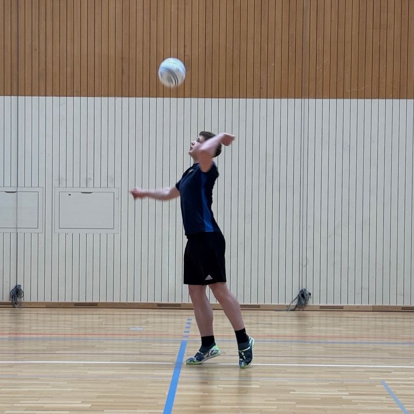 Ein Mann befindet sich in einer Halle, springt und wirft einen Ball. Der Platz hat Holzböden und weiße Wände mit braunen Paneelen.