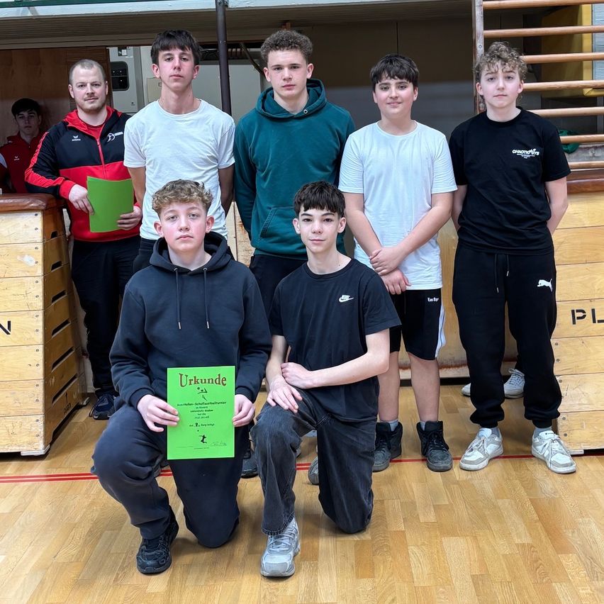 Sieben junge Männer posieren für ein Foto in einer Turnhalle. Einer hält ein Zertifikat, andere stehen hinter ihm und tragen Sneaker und Kapuzenpullover. Holzkisten und eine rote Linie auf dem Boden sind sichtbar.