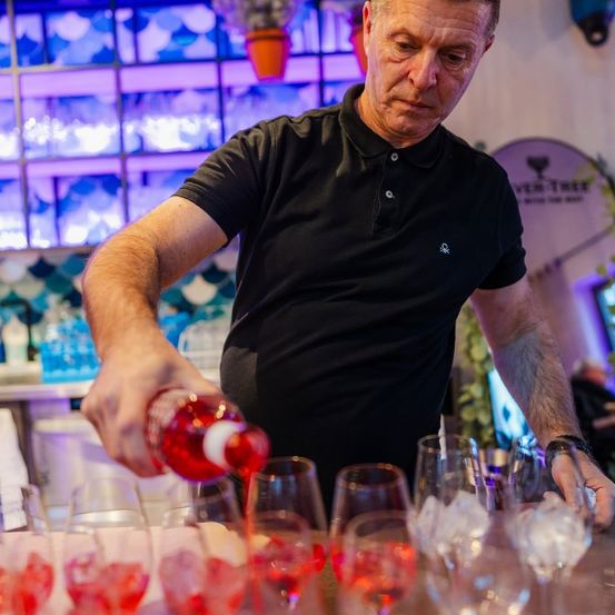 Ein Barkeeper gießt rote Flüssigkeit in Weingläser in einer Bar mit bunten Lichtern und einer dekorativen Wand.
