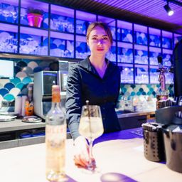 Eine Frau steht hinter einer Bartheke und hält ein Glas Wein und eine Flasche Schnaps. Die Bar ist mit lila Lichtern beleuchtet, und verschiedene Küchengeräte sind im Hintergrund zu sehen.