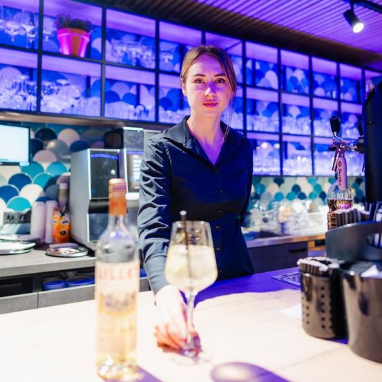 Eine Frau steht hinter einer Bartheke und hält ein Glas Wein und eine Flasche Schnaps. Die Bar ist mit lila Lichtern beleuchtet, und verschiedene Küchengeräte sind im Hintergrund zu sehen.