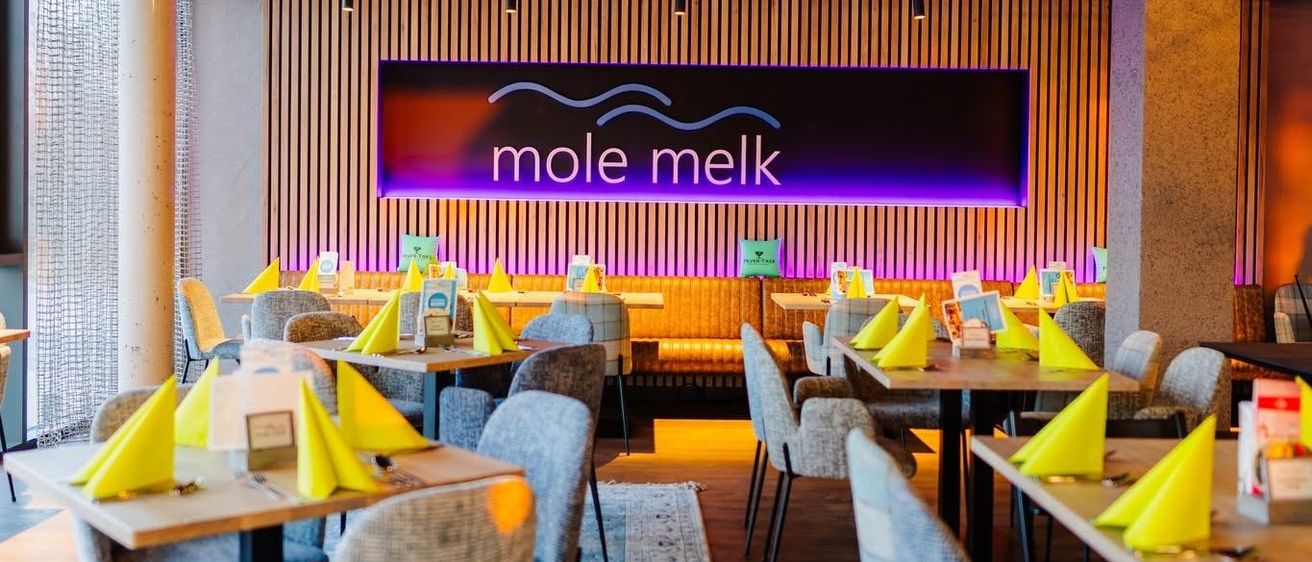 Ein Restaurant mit modernem Design, mit Holzböden, Tischen mit gelben Servietten und einem großen Schild mit der Aufschrift 'mole melk' in lila Beleuchtung.