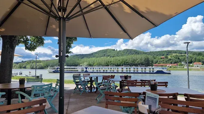 Ein Außenrestaurant mit Tischen und Stühlen unter Sonnenschirmen, mit Blick auf einen Fluss und ein Kreuzfahrtschiff in der Ferne.