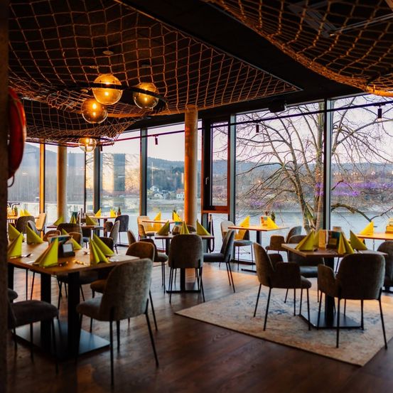 Ein Restaurant mit großen Fenstern, das einen malerischen Blick auf einen Fluss und Berge bietet. Die Tische sind mit gelben Servietten gedeckt und der Raum ist von warmen Lichtern erhellt.