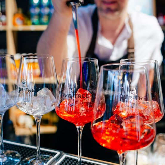 Ein Barkeeper gießt rote Flüssigkeit in Weingläser mit Eiswürfeln, hinter einer Bartheke stehend.