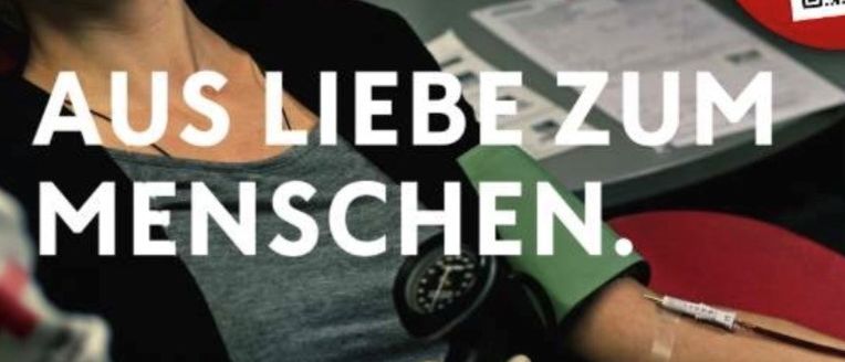 Eine Frau lächelt, während ihr der Blutdruck gemessen wird. Auf dem Schreibtisch befindet sich ein Blutspende-Schild.