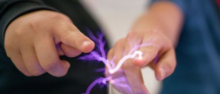 Zwei Menschen experimentieren mit einem violetten elektrischen Bogen zwischen ihren Fingerspitzen. Einer trägt Schwarz, der andere Blau.