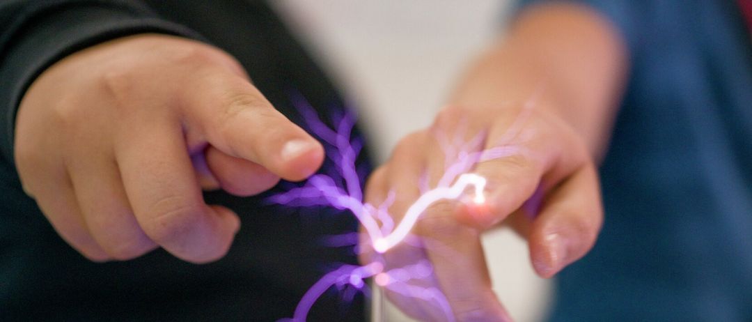 Zwei Menschen experimentieren mit einem violetten elektrischen Bogen zwischen ihren Fingerspitzen. Einer trägt Schwarz, der andere Blau.